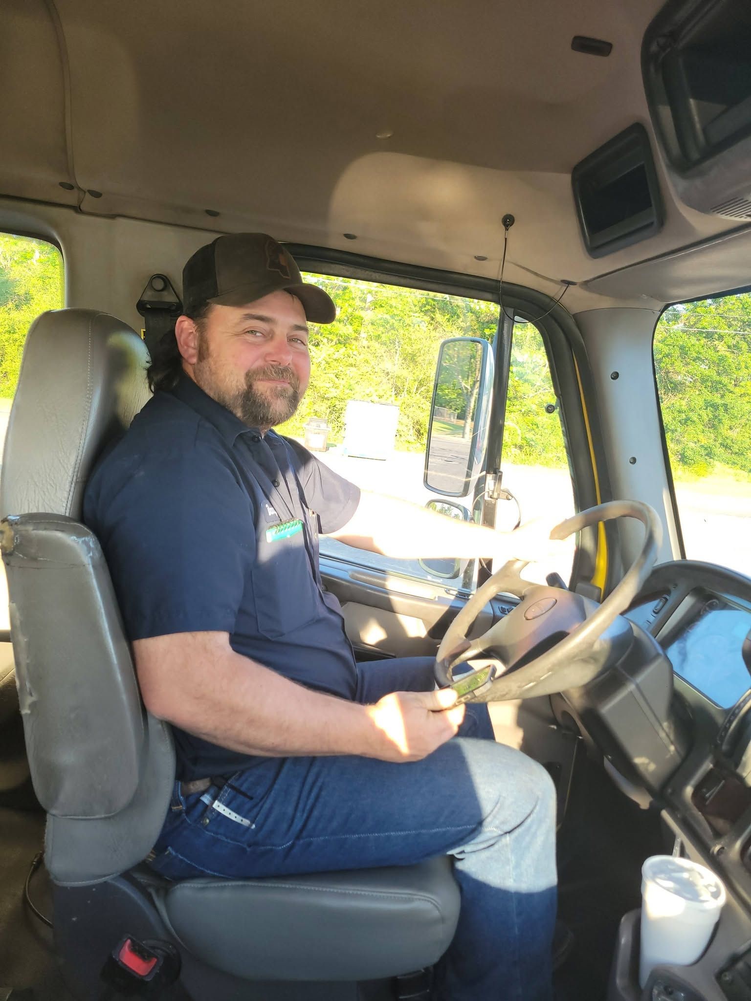 Man sitting in the driver’s seat of a vehicle, wearing a black cap and shirt, looking toward the camera.