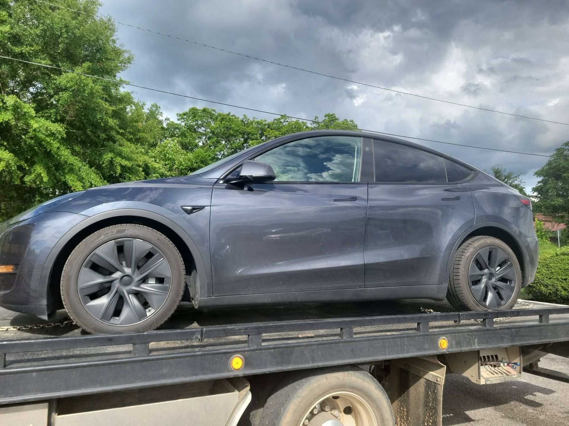 Dark gray sedan on a flatbed trailer outdoors near trees under a cloudy sky