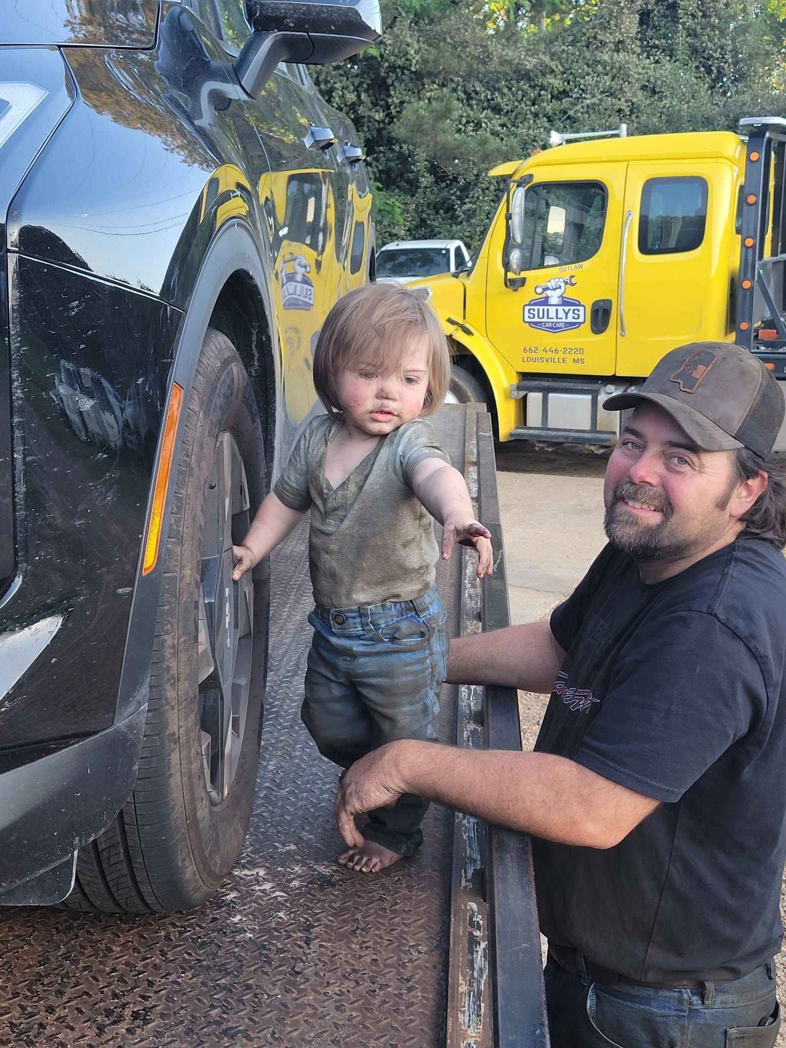 Child standing by a large truck tire while a man steadies them; yellow truck in the background.