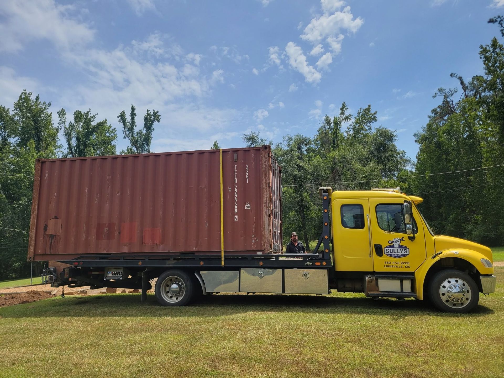 Yellow semi-truck hauling a red shipping container on a grassy road with trees and blue sky.