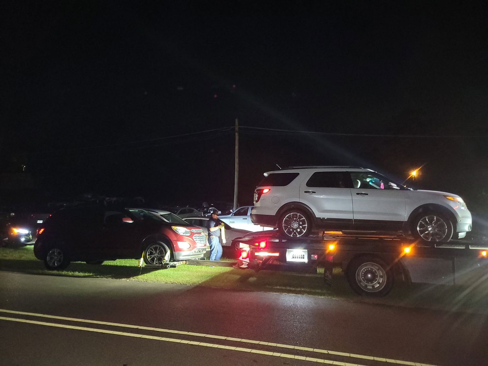 Tow truck hauling a white SUV at night, with a black car and flashing lights on the roadside