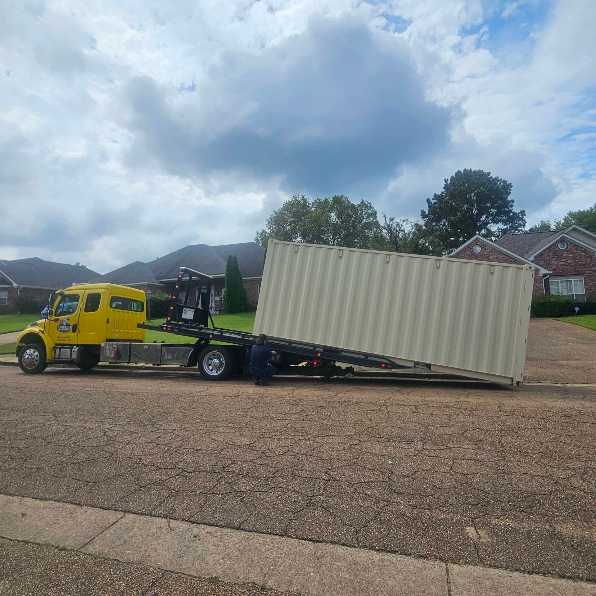 Yellow tow truck hauling a tilted beige shipping container on a lowboy trailer in a suburban lot under cloudy skies