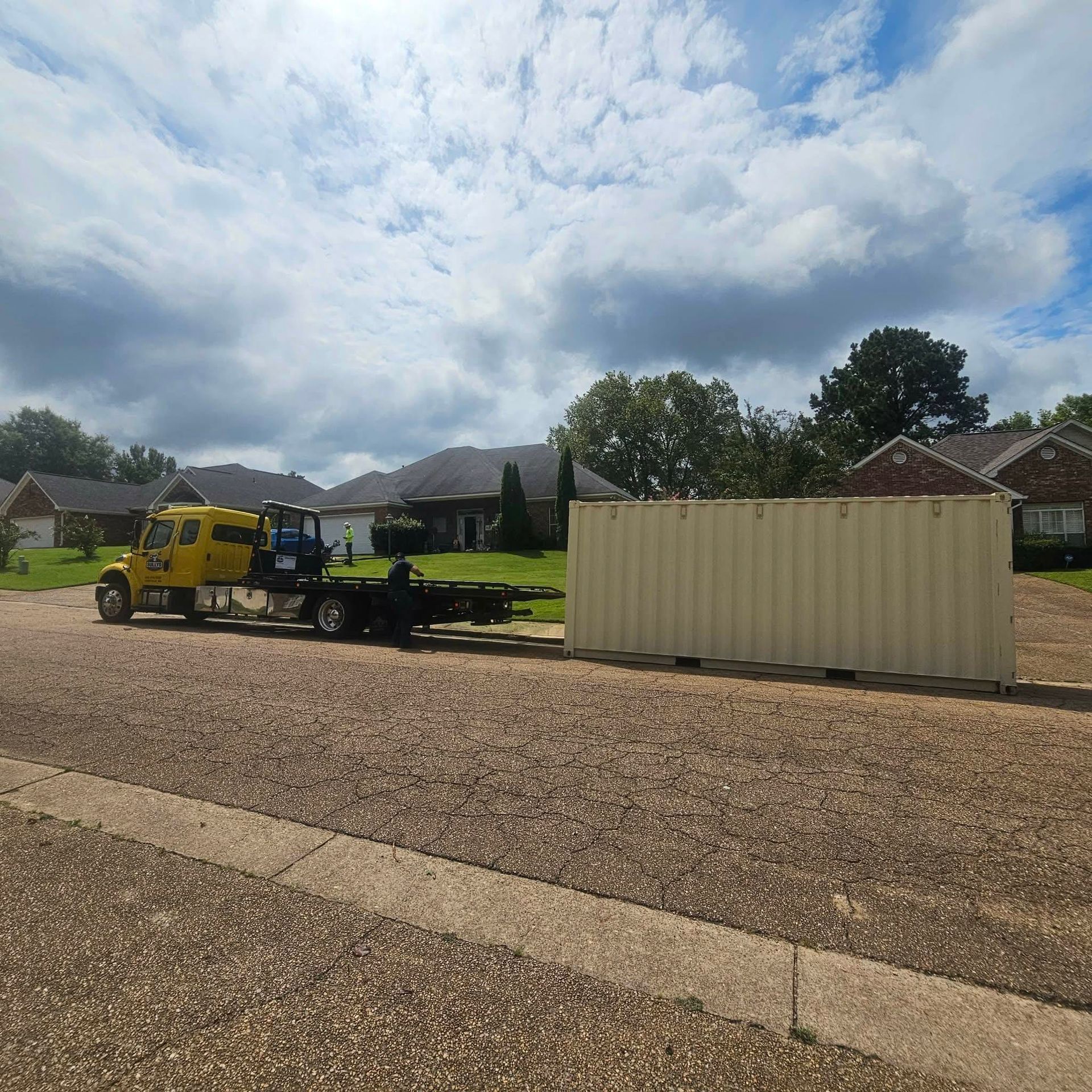 Yellow truck hauling a beige shipping container in a suburban gravel lot under a cloudy sky