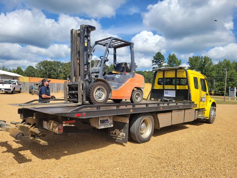 Orange forklift on a flatbed tow truck in a gravel lot under a cloudy sky