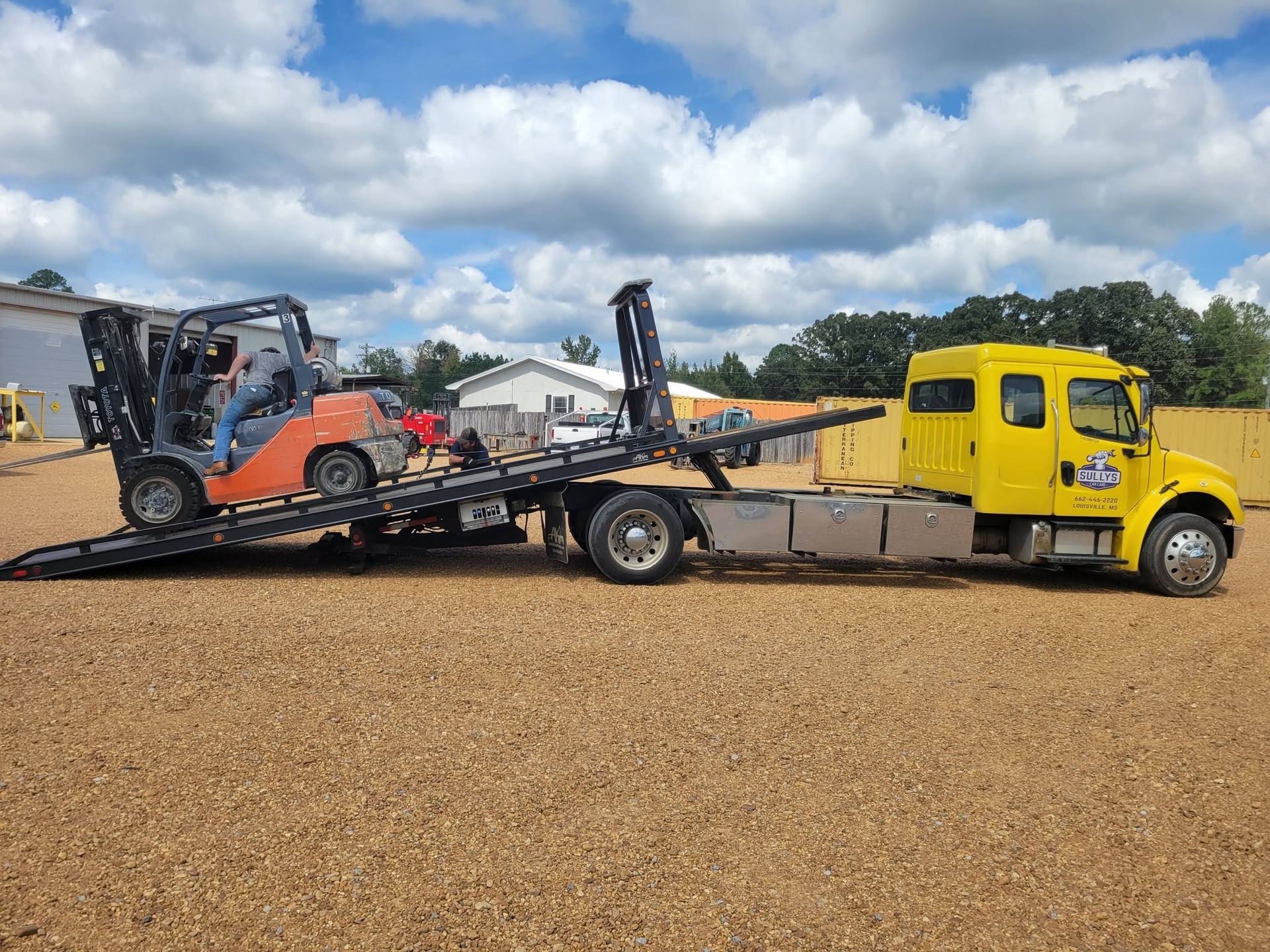 Yellow tow truck carrying a forklift on a flatbed trailer in a gravel lot under cloudy skies