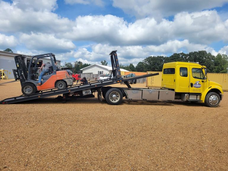 Yellow tow truck carrying a skid steer on a flatbed in a gravel lot under a cloudy sky