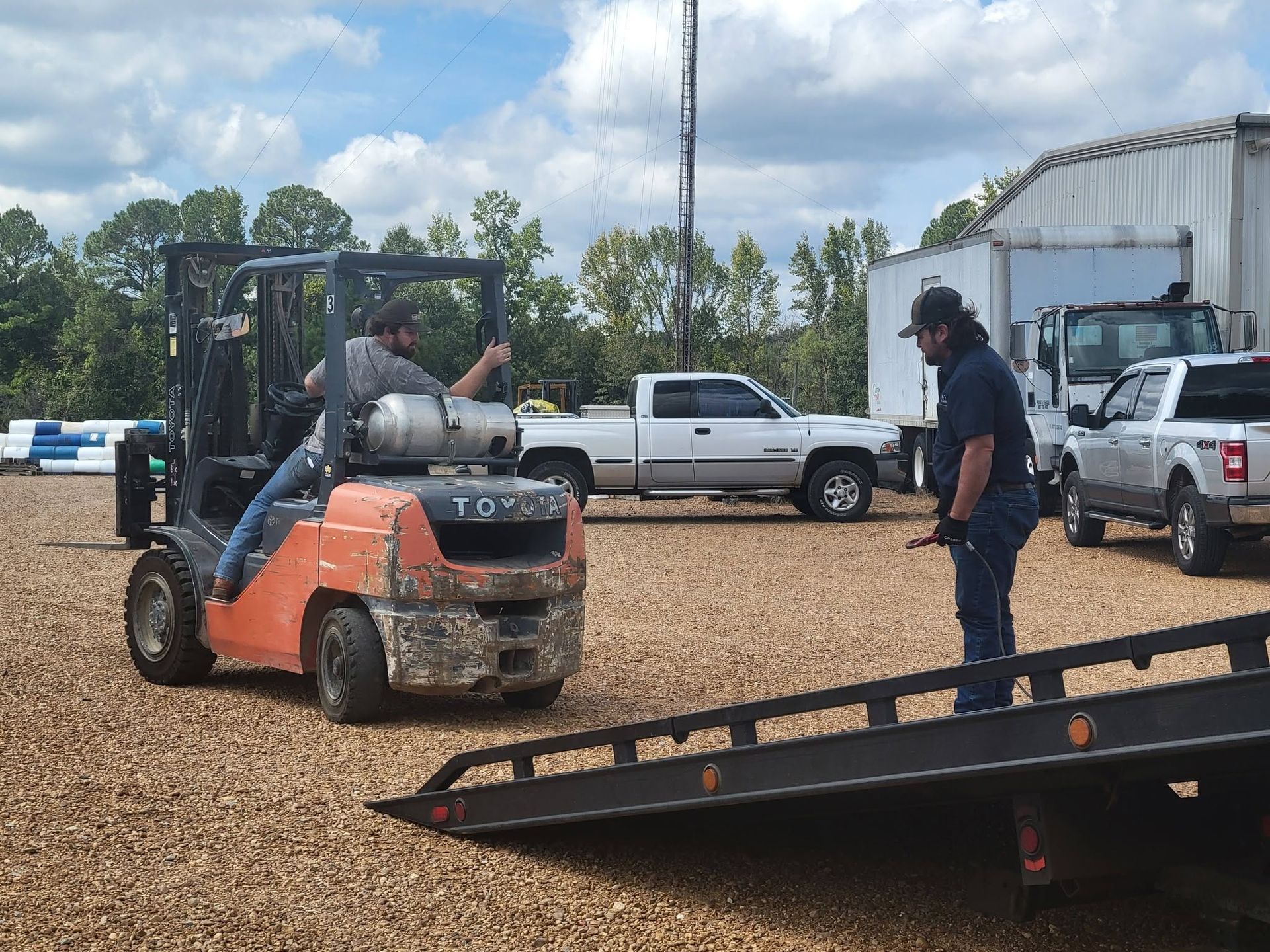 Forklift loading equipment onto a flatbed trailer in a gravel yard, with workers nearby.