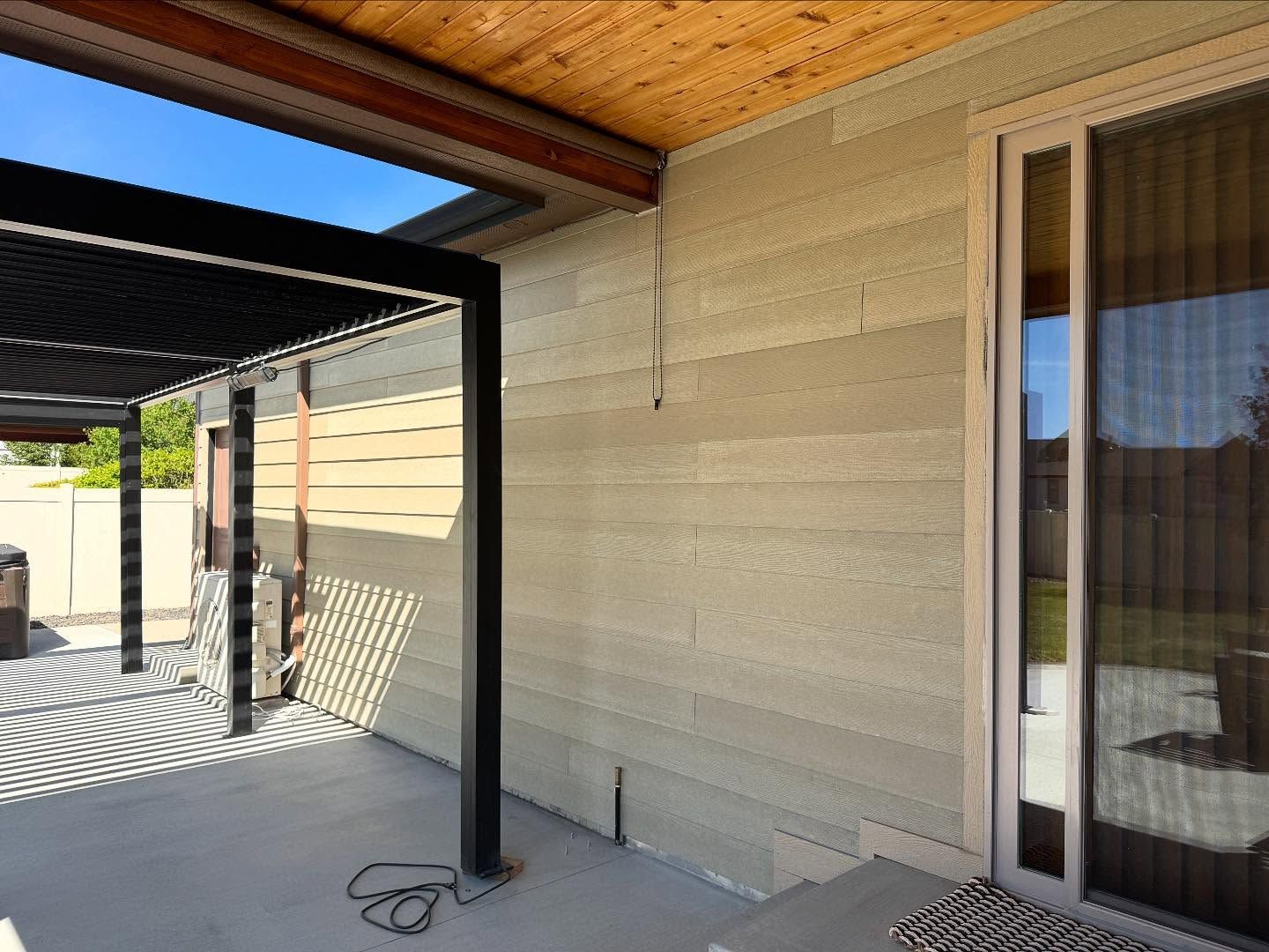 A patio featuring a wooden ceiling, a black pergola, a sliding glass door, and a textured, layered light-tan earth wall.