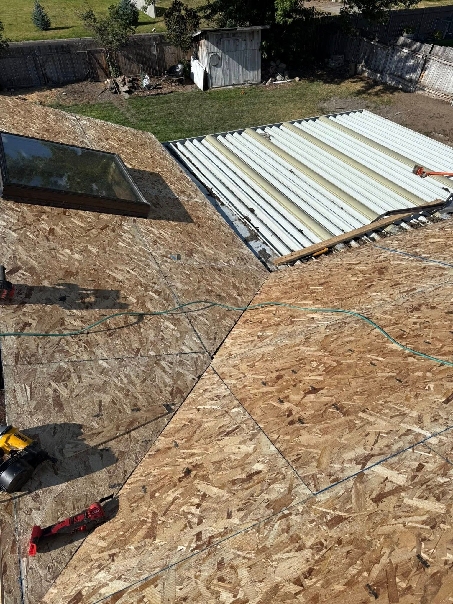 A roof under construction with wooden plywood decking, a skylight, and a section of exposed white corrugated roofing.