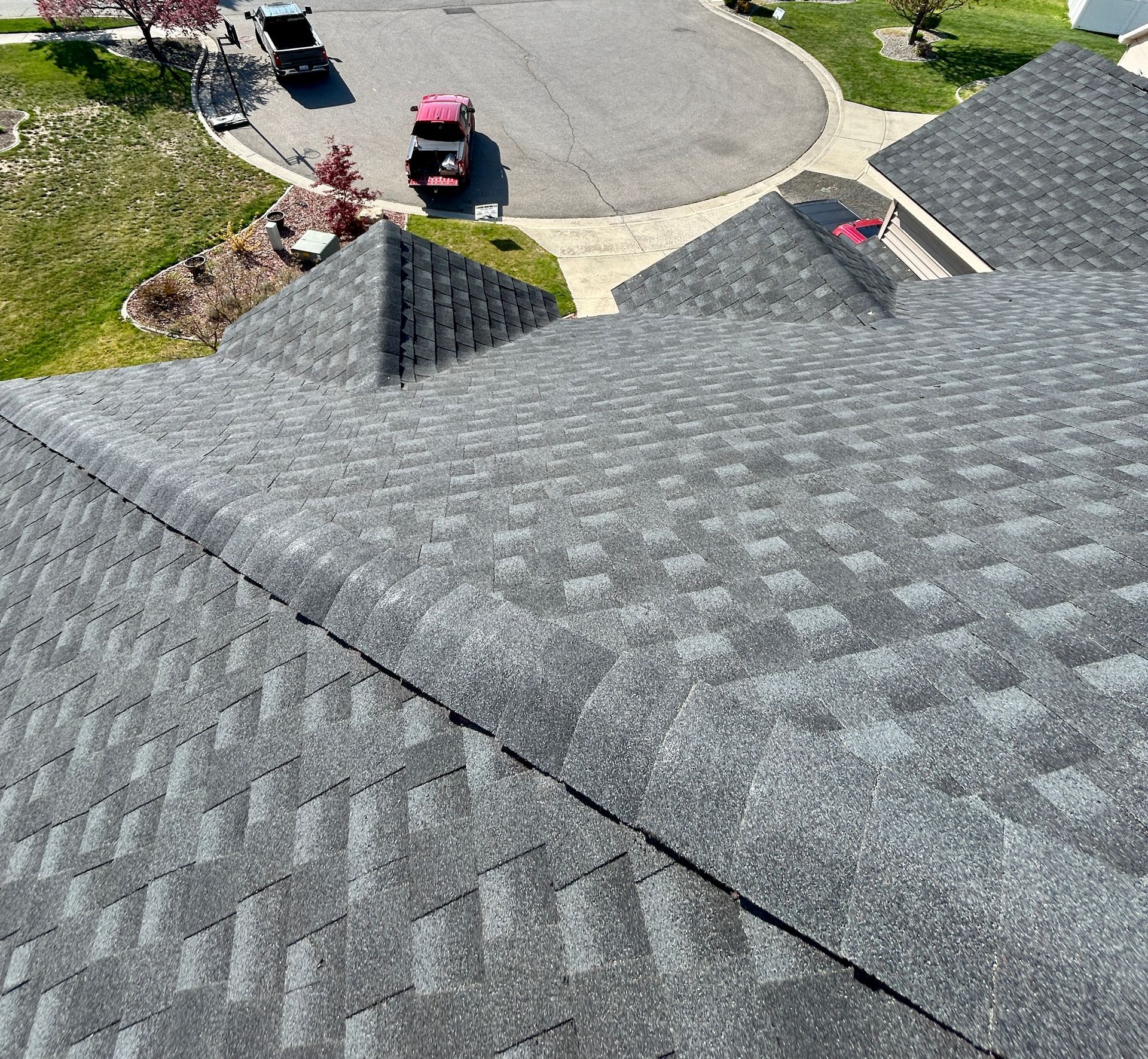 High-angle view of a dark gray shingled roof, looking down toward a suburban driveway with two parked vehicles.