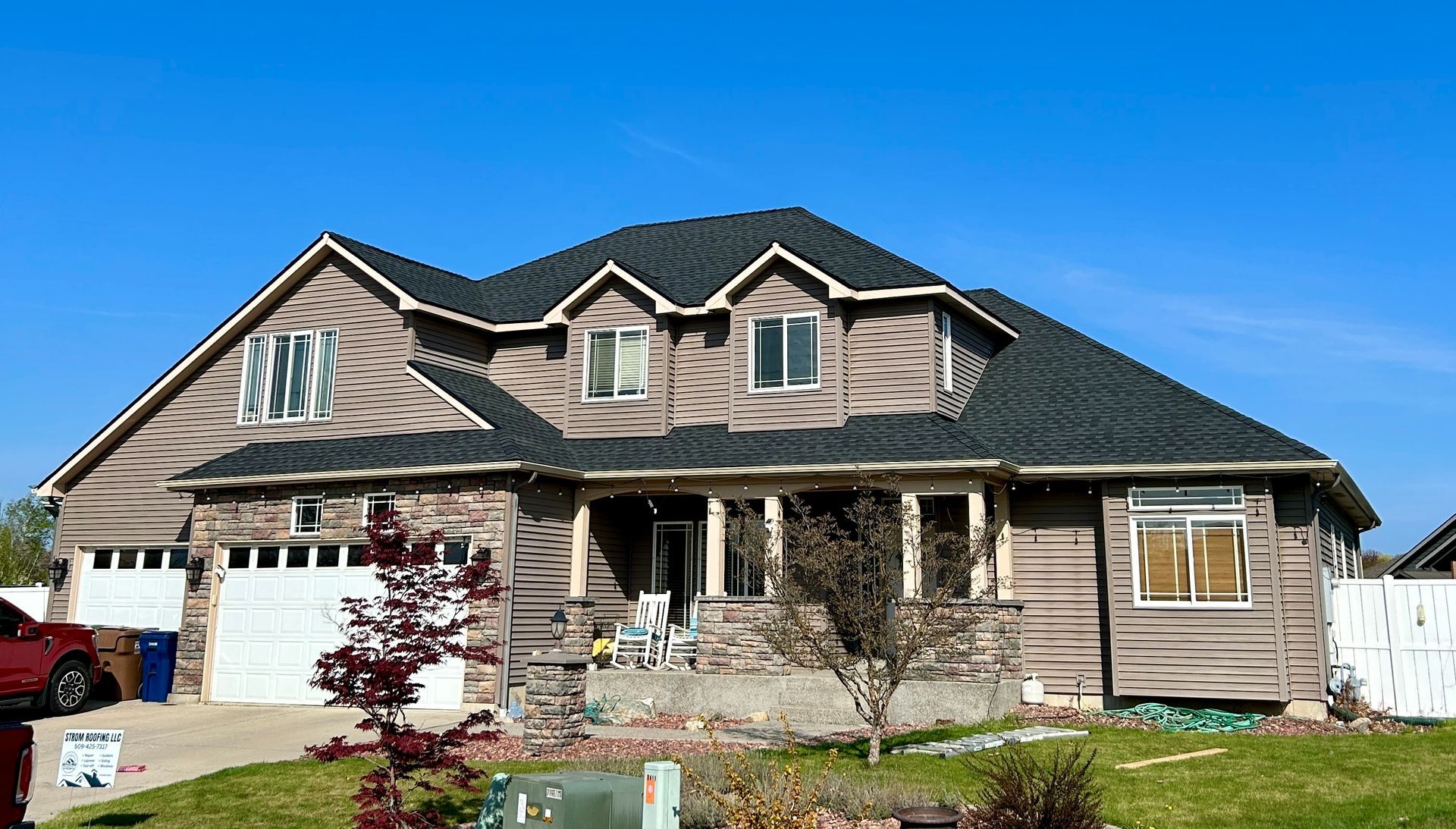 A large, multi-story tan house with a dark roof, two garage doors, and a covered front porch under a bright blue sky.