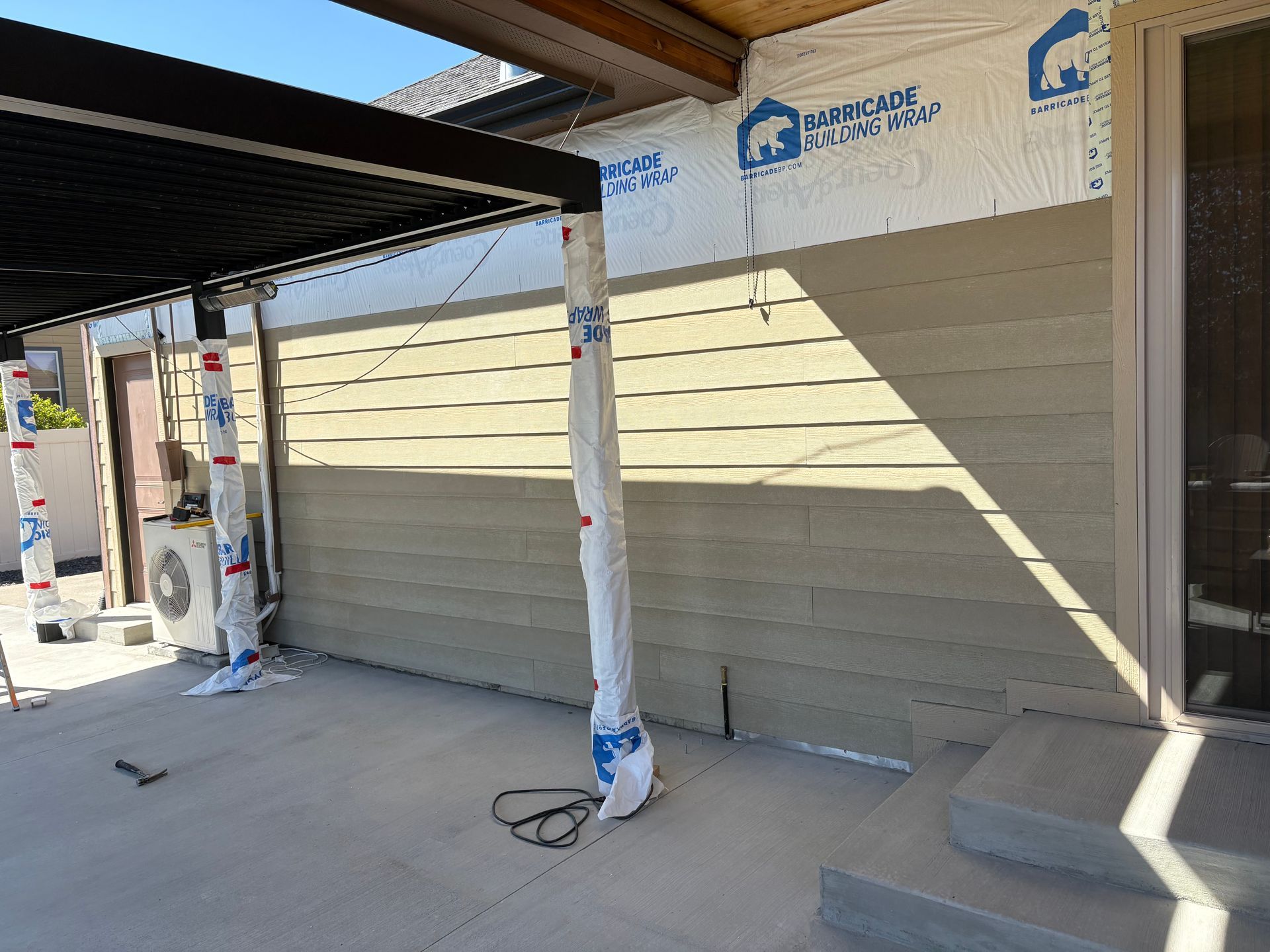 Tan siding with exposed white house wrap and black patio posts under construction on a concrete patio.