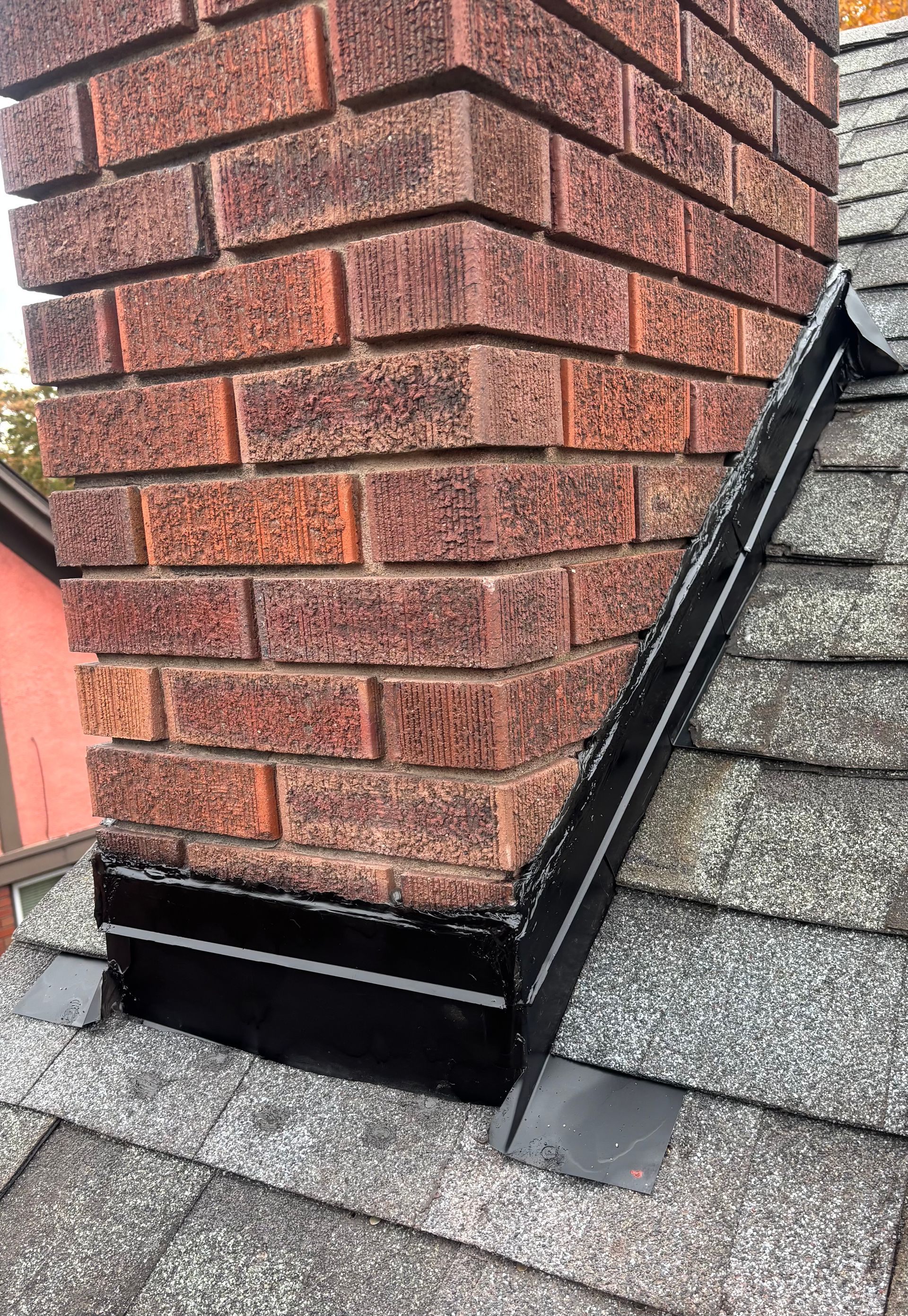 Black metal chimney flashing installed against red brick and grey asphalt roof shingles.