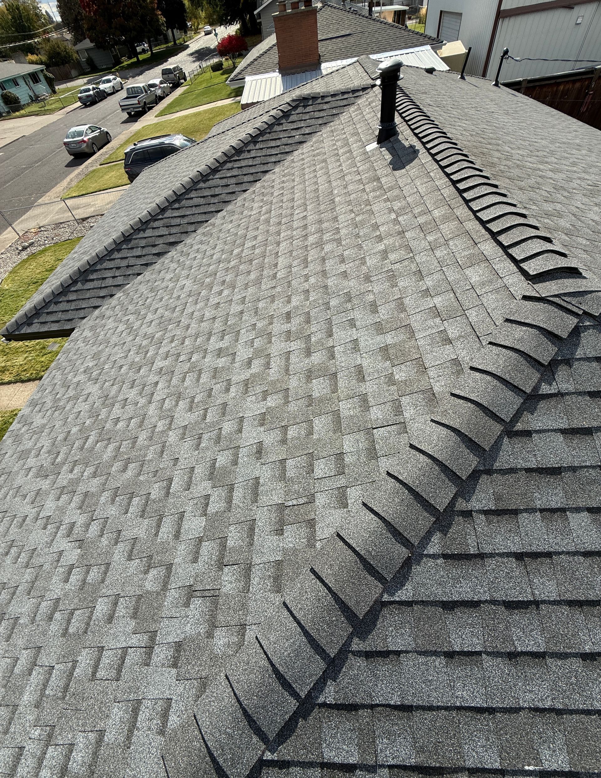 A high-angle view of a gray asphalt shingle roof with prominent ridges, a chimney, and a suburban street in the background.