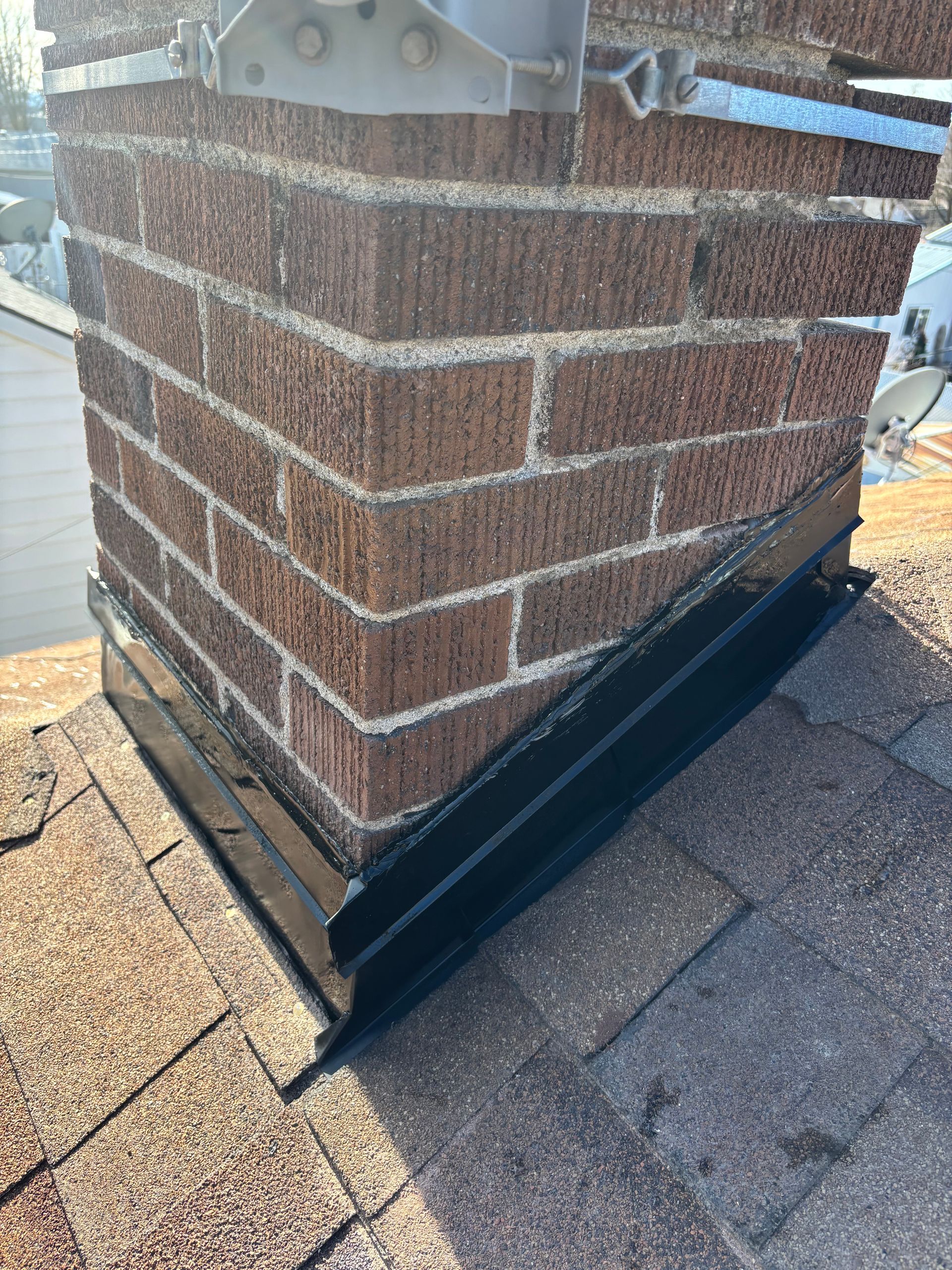 A close-up view of dark metal flashing installed around a brick chimney on a shingled roof.
