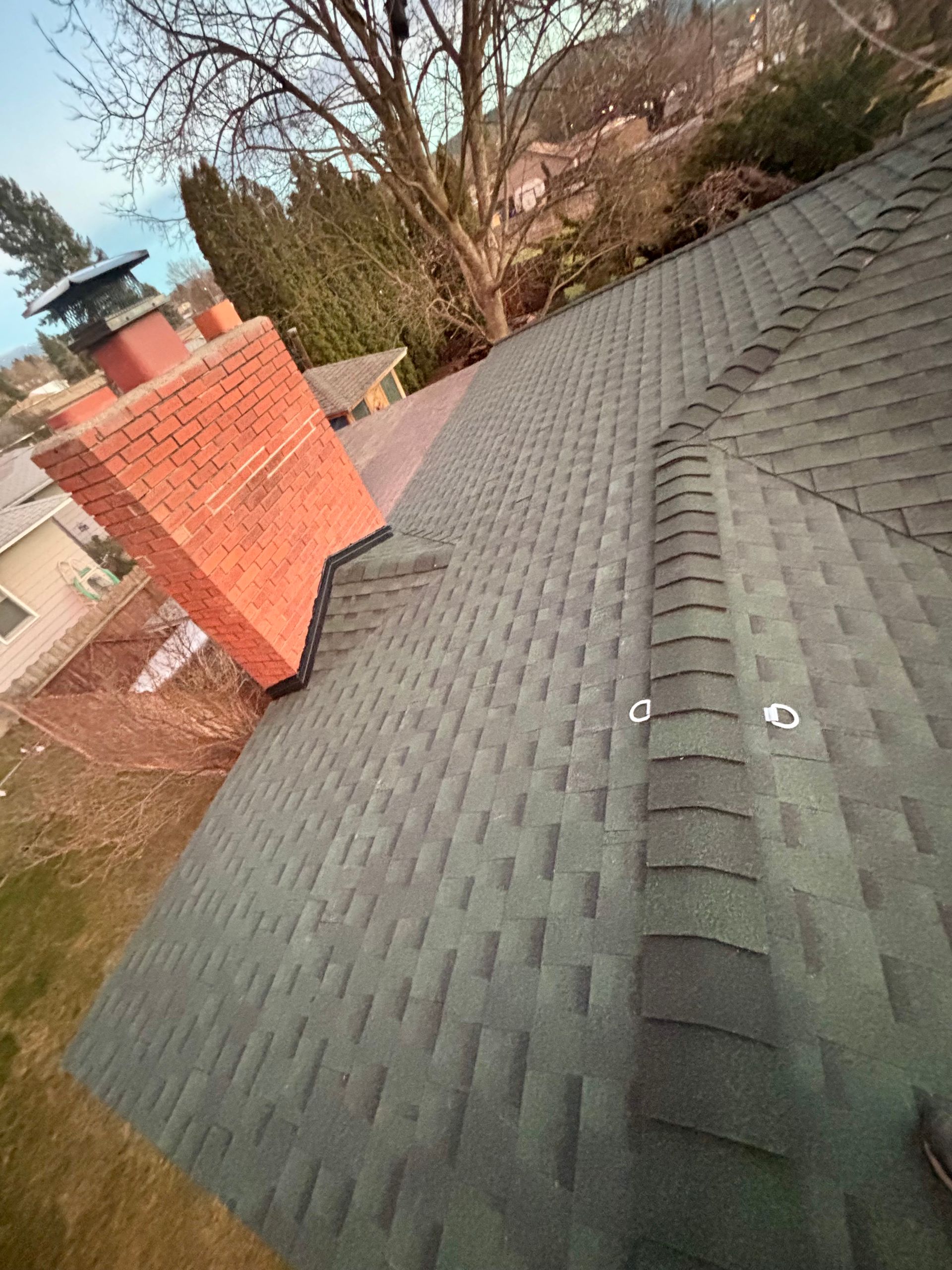 A high-angle view of a dark shingled roof featuring a red brick chimney and two metal roof anchors on the ridge line.