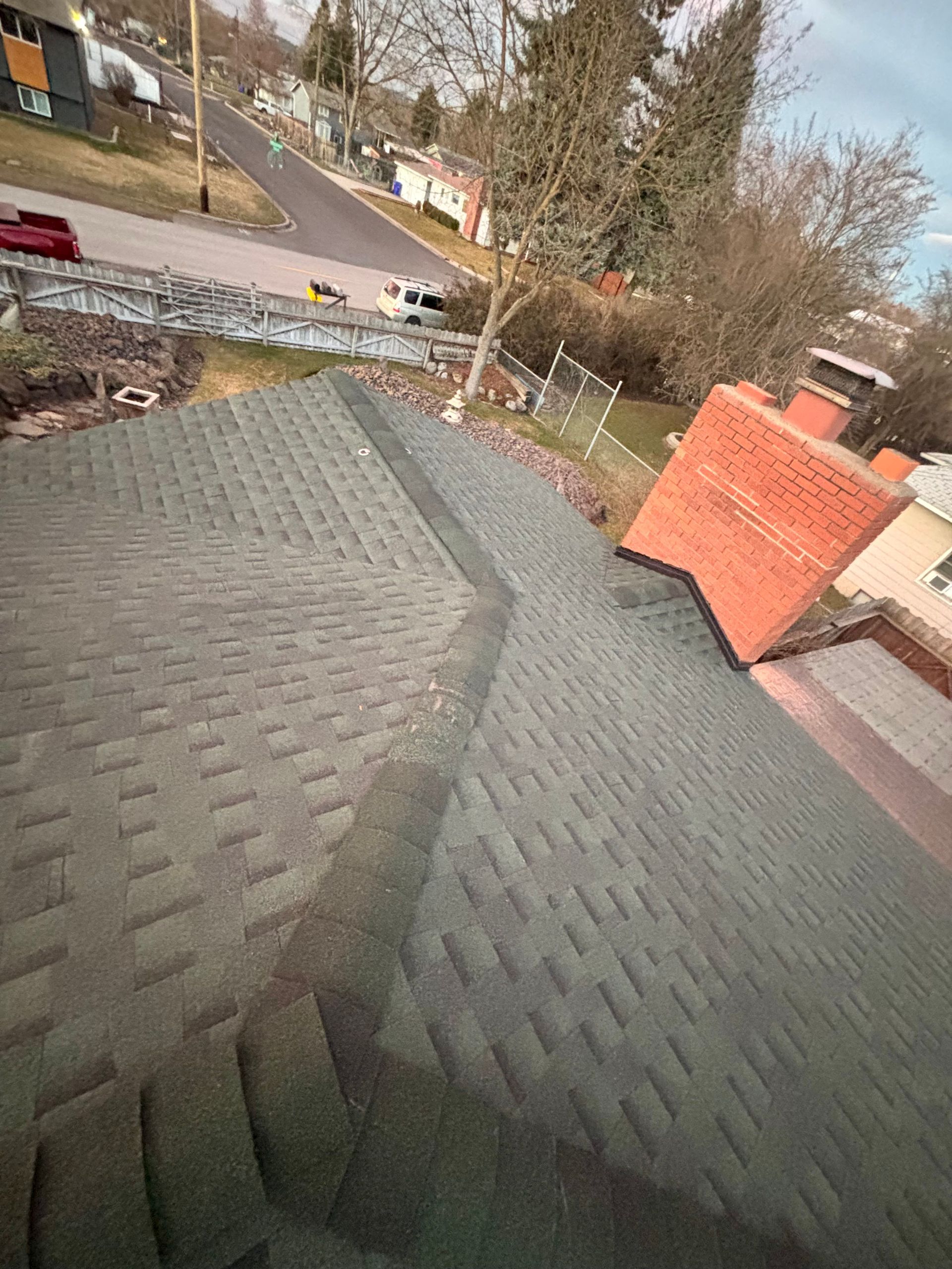 A high-angle view looking down at a dark gray shingled roof with a red brick chimney and a nearby street.