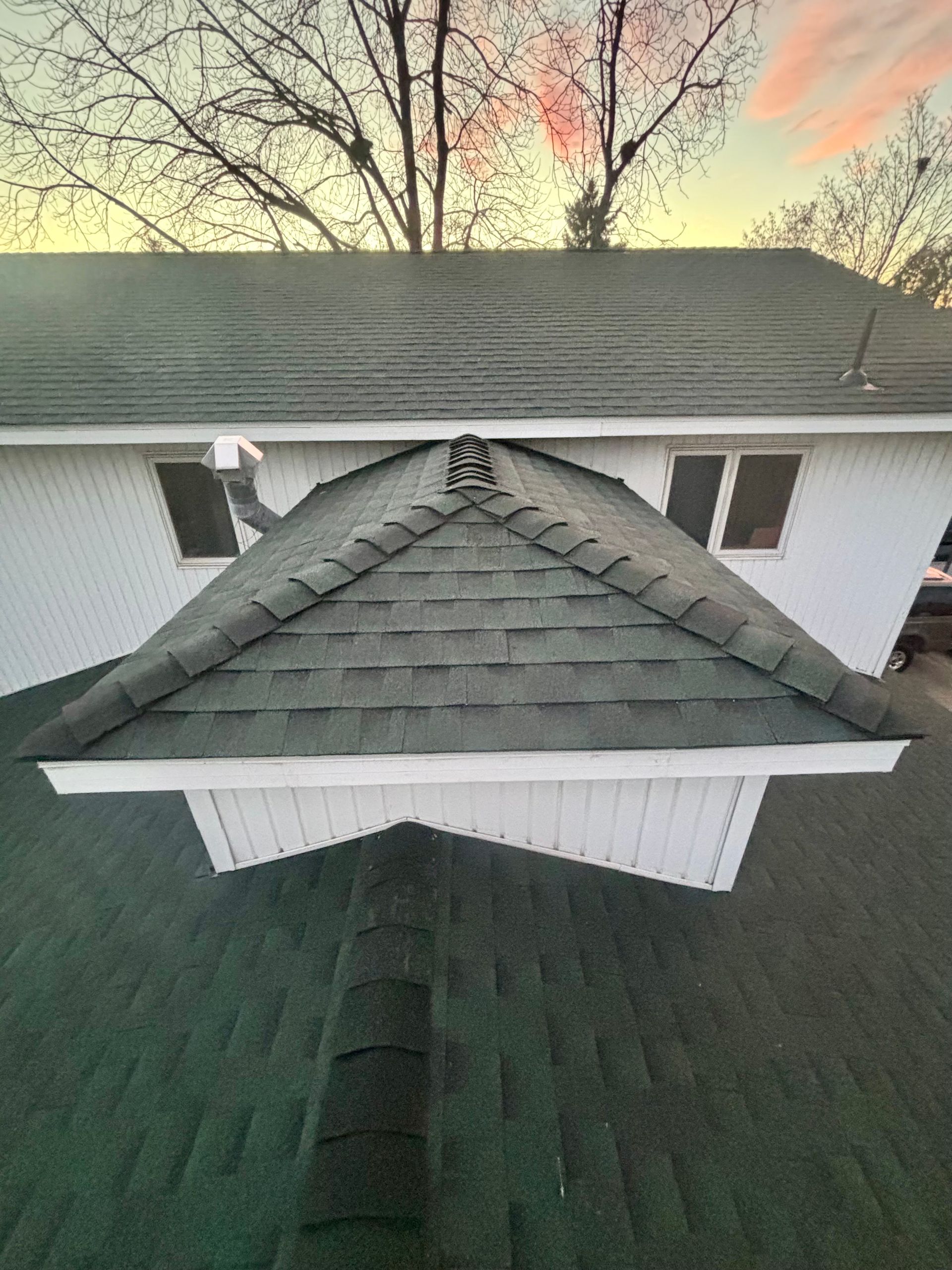 A high-angle view of a dark shingled roof with a pyramidal dormer, overlooking a larger house section at sunset.