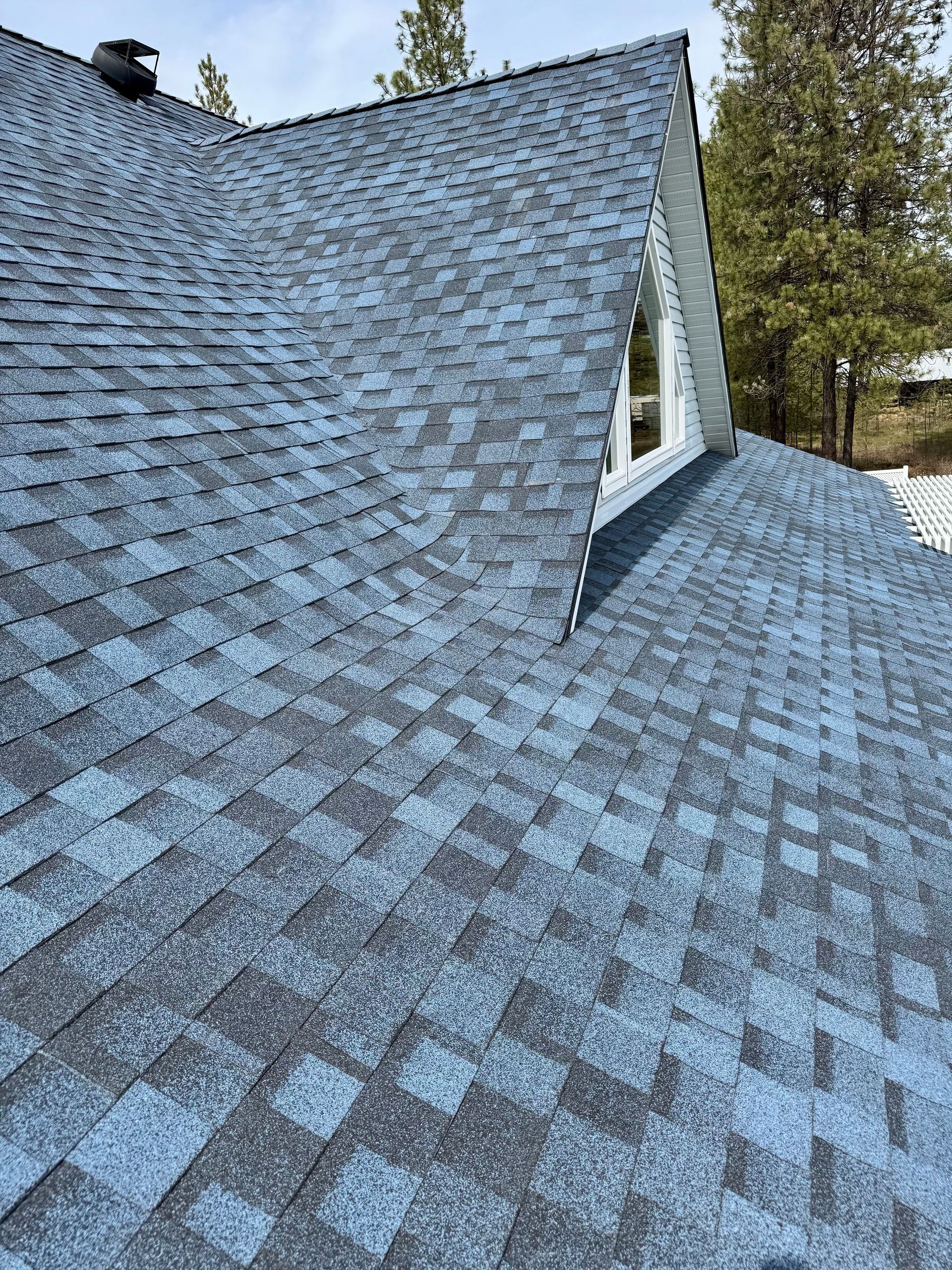 A close-up view of a blue and grey shingled roof with a small dormer window and pine trees in the background.