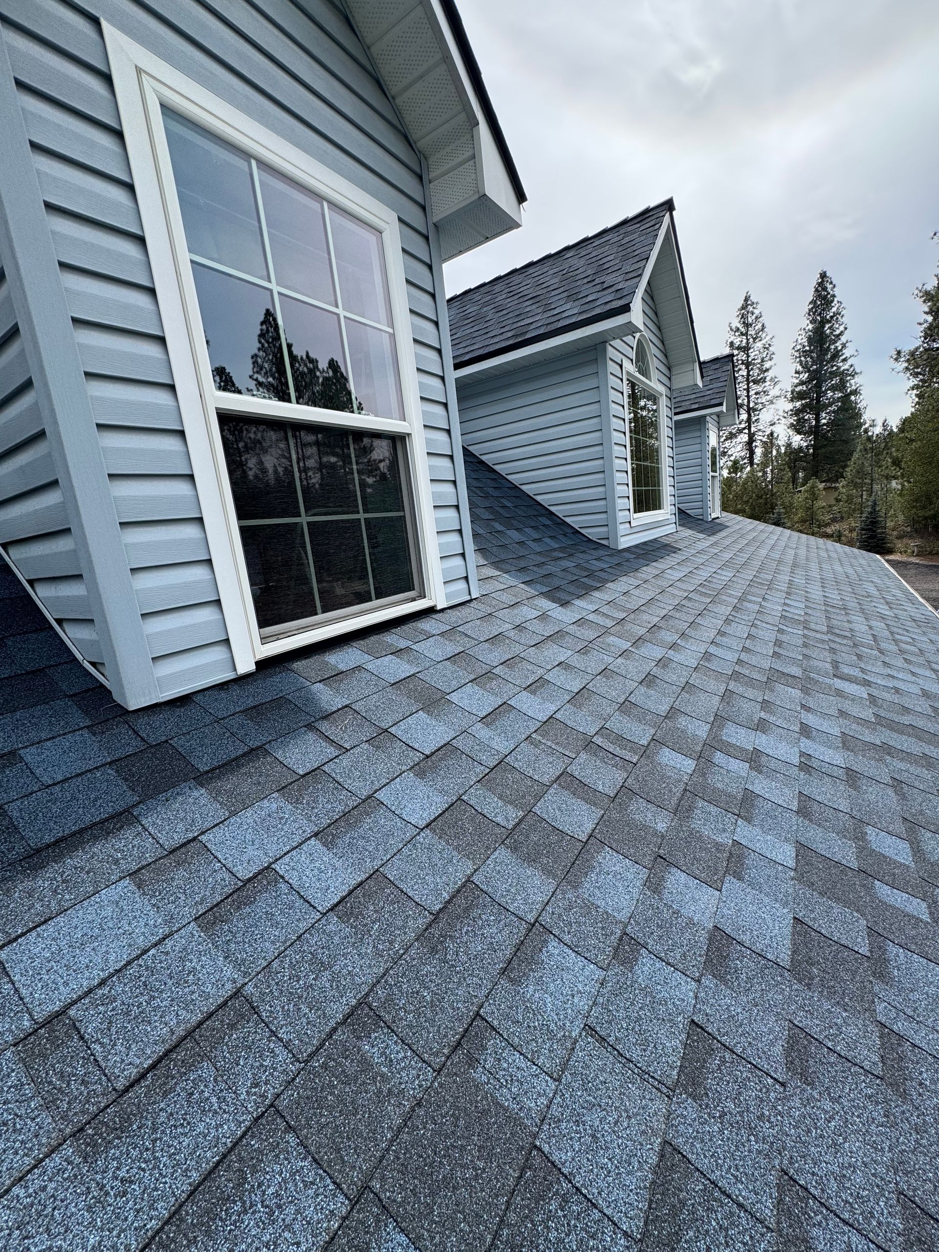 A view of a blue shingled residential roof with two dormer windows and light blue siding under a cloudy sky.
