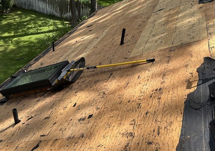 An angled view of a residential roof undergoing repair, showing exposed plywood decking, plumbing vents, and a skylight.