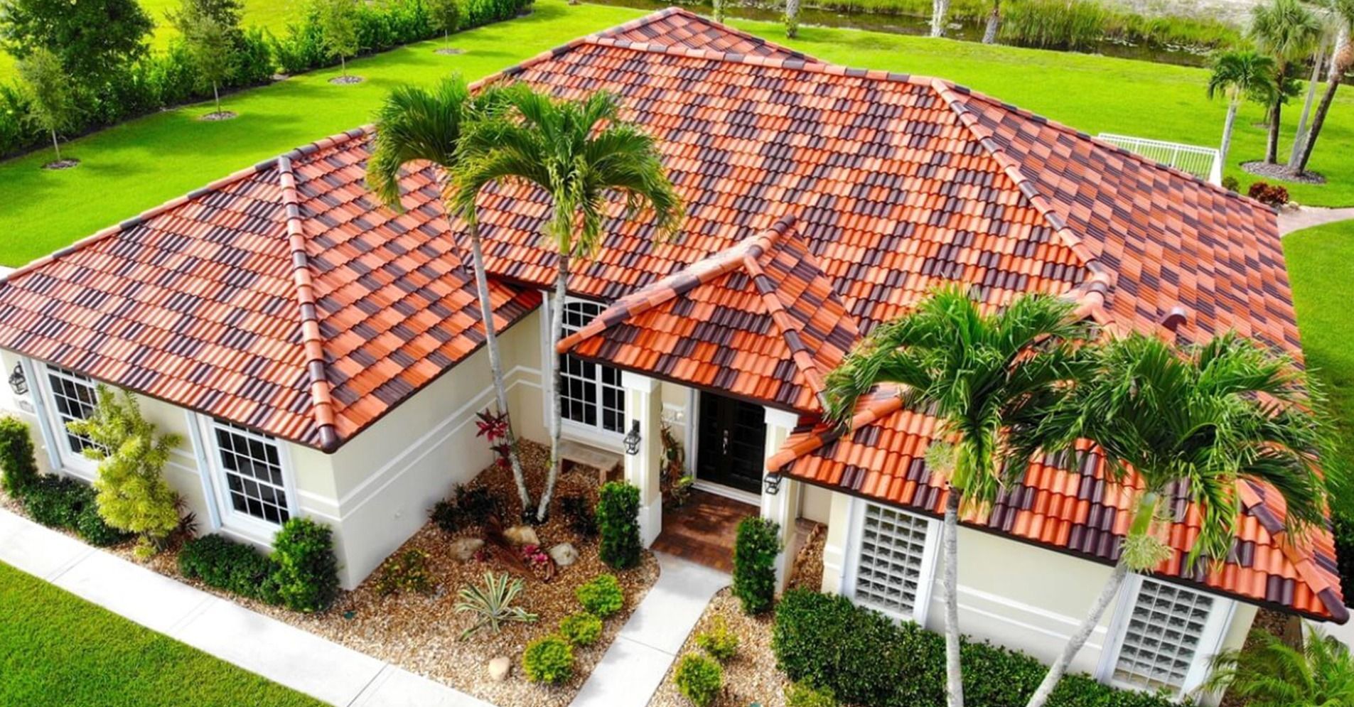 An aerial view of a single-story house with a red and black patterned tile roof, white stucco walls, and palm trees.