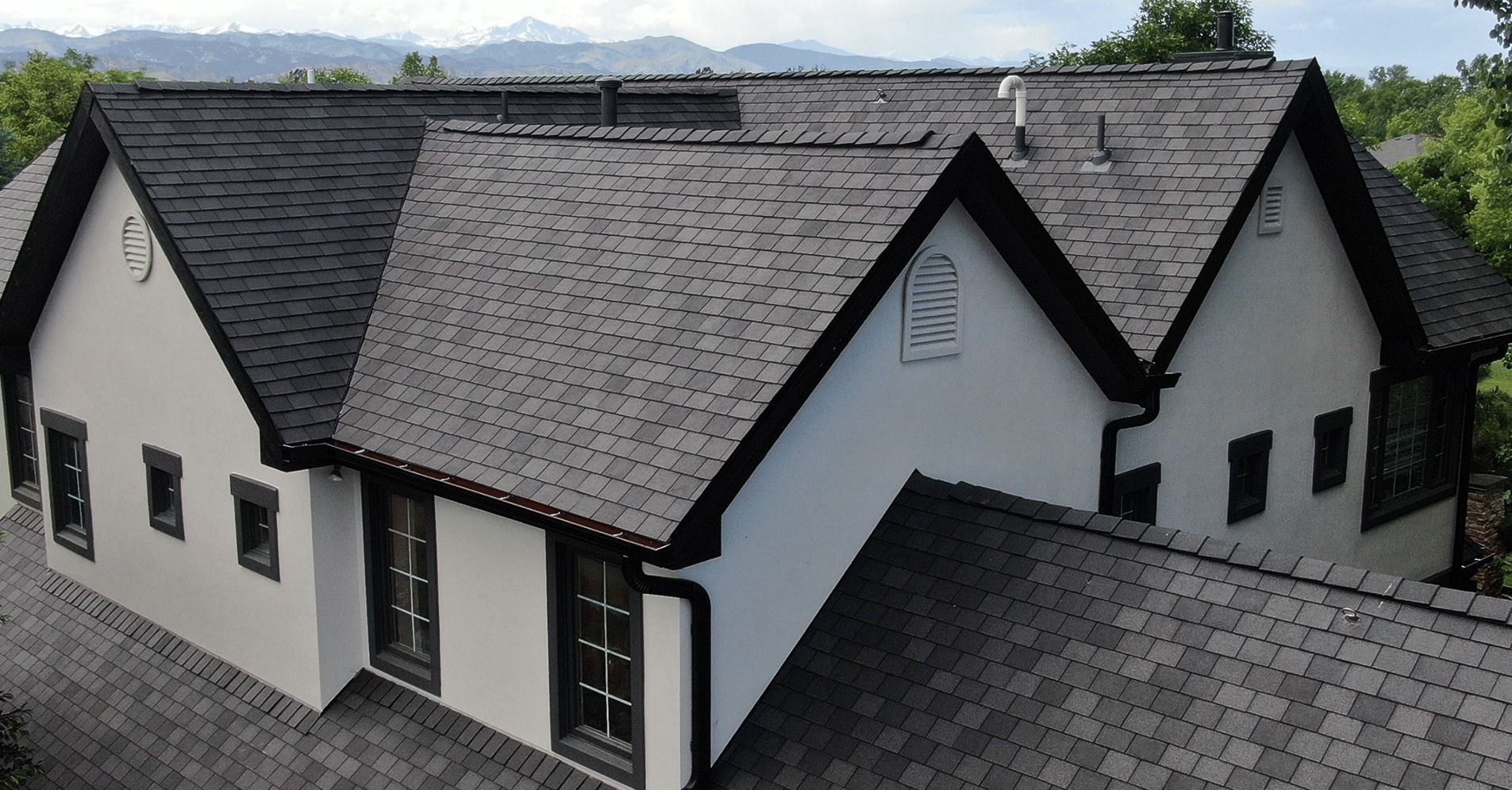 High-angle view of a multi-gabled house with grey shingles, cream-colored stucco walls, and black trim.