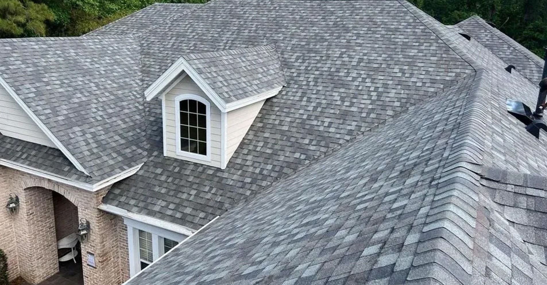 A high-angle view of a gray shingled roof on a house with a gabled dormer and beige brick siding.