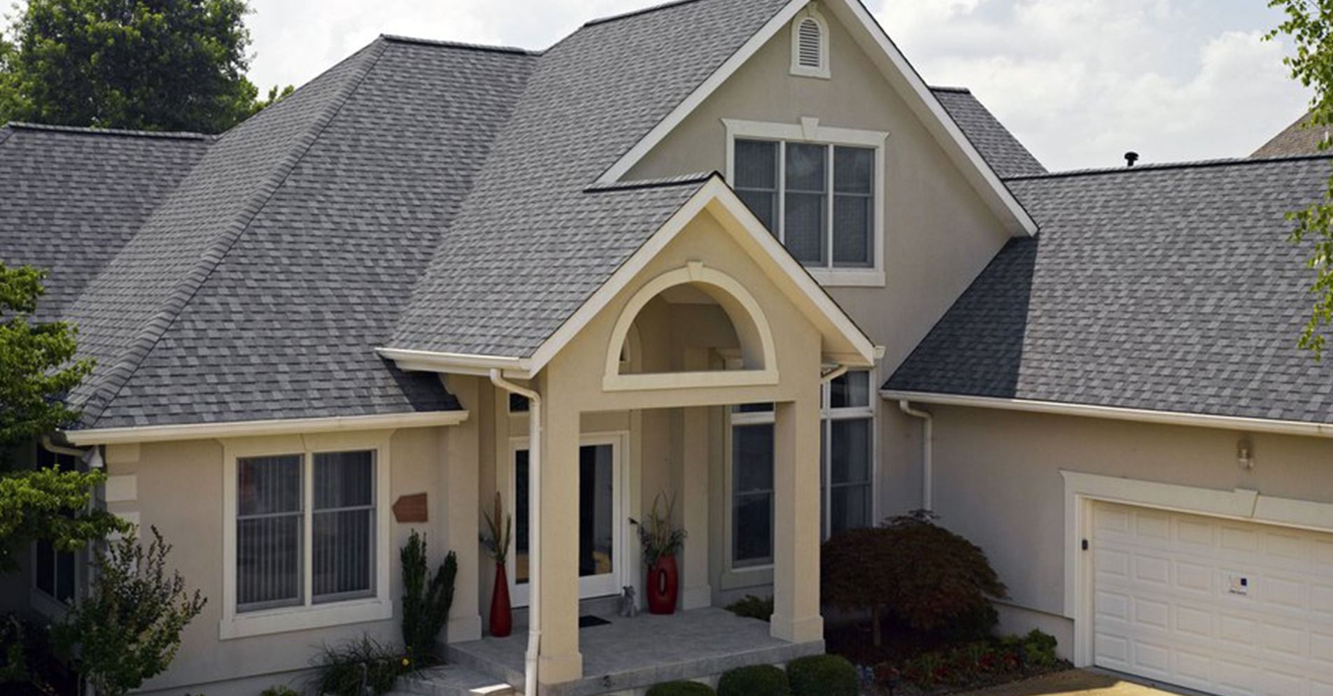 A two-story tan suburban house with a gray shingled roof, a front porch entry with an arch, and an attached garage.