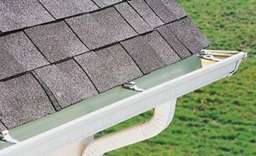 A white metal gutter attached to a roof edge with shingles above and a downspout below against a grassy background.