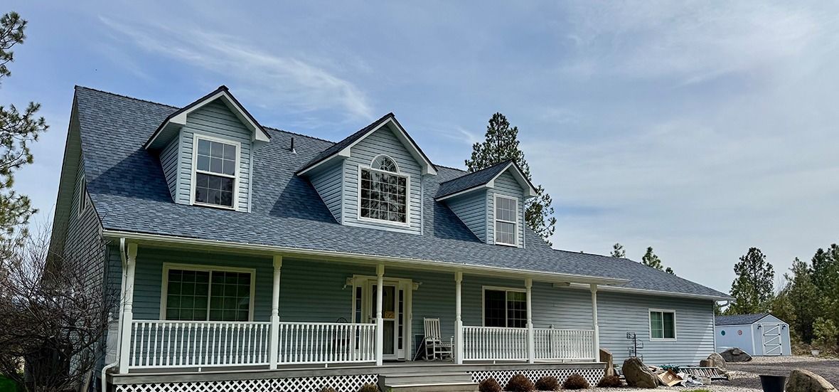 A two-story light blue house with a covered front porch, three gabled dormers on the roof, and a pale blue sky background.