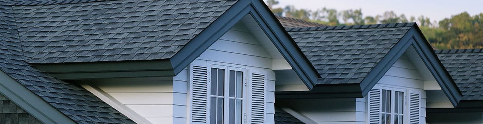 A close-up of house dormers with dark gray shingles and white siding against a blurred, hilly background.