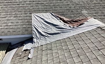 A large, grey tarp is draped over a section of grey asphalt shingles on a sloped roof to cover damaged areas.