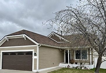 Tan single-story house with a brown garage door, dark roof, and a small front porch under a cloudy sky.