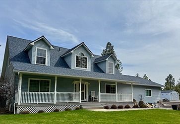 A two-story, light blue house with a wide front porch, white railings, and three gabled dormers under a blue sky.