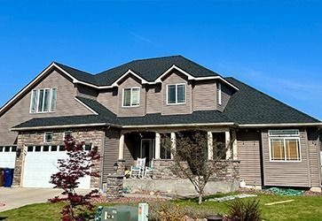 Two-story house with dark shingles, brown siding, stone accents, a two-car garage, and a front porch under a clear sky.