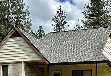 An A-frame house exterior featuring light yellow siding, a brick section, and a dark-shingled roof under a cloudy sky.