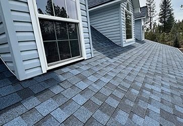 A close-up of a blue and grey asphalt shingle roof, with light blue siding and windows of dormers visible above.