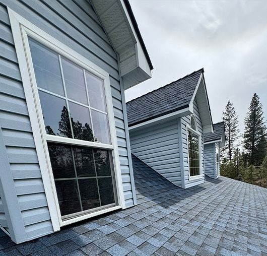 The exterior of a light blue house with multiple dormer windows and dark gray shingled roofs against a cloudy sky.