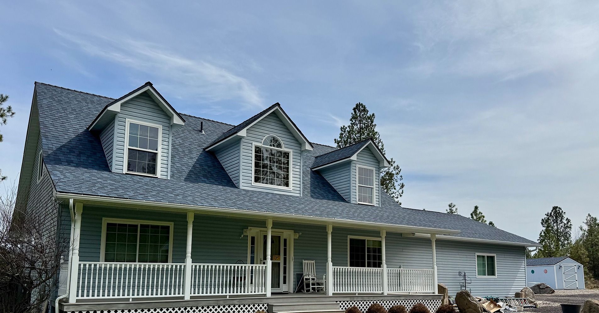 A light blue house with a covered front porch, dormer windows, and a dark, patterned roof under a blue sky.