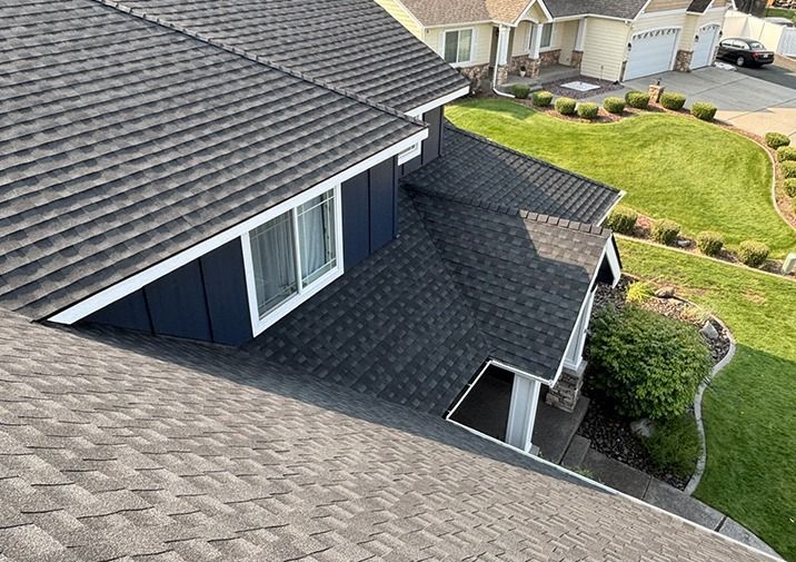 An elevated, angled view of a house roof with dark gray shingles, a blue-sided dormer with a window, and a suburban lawn.