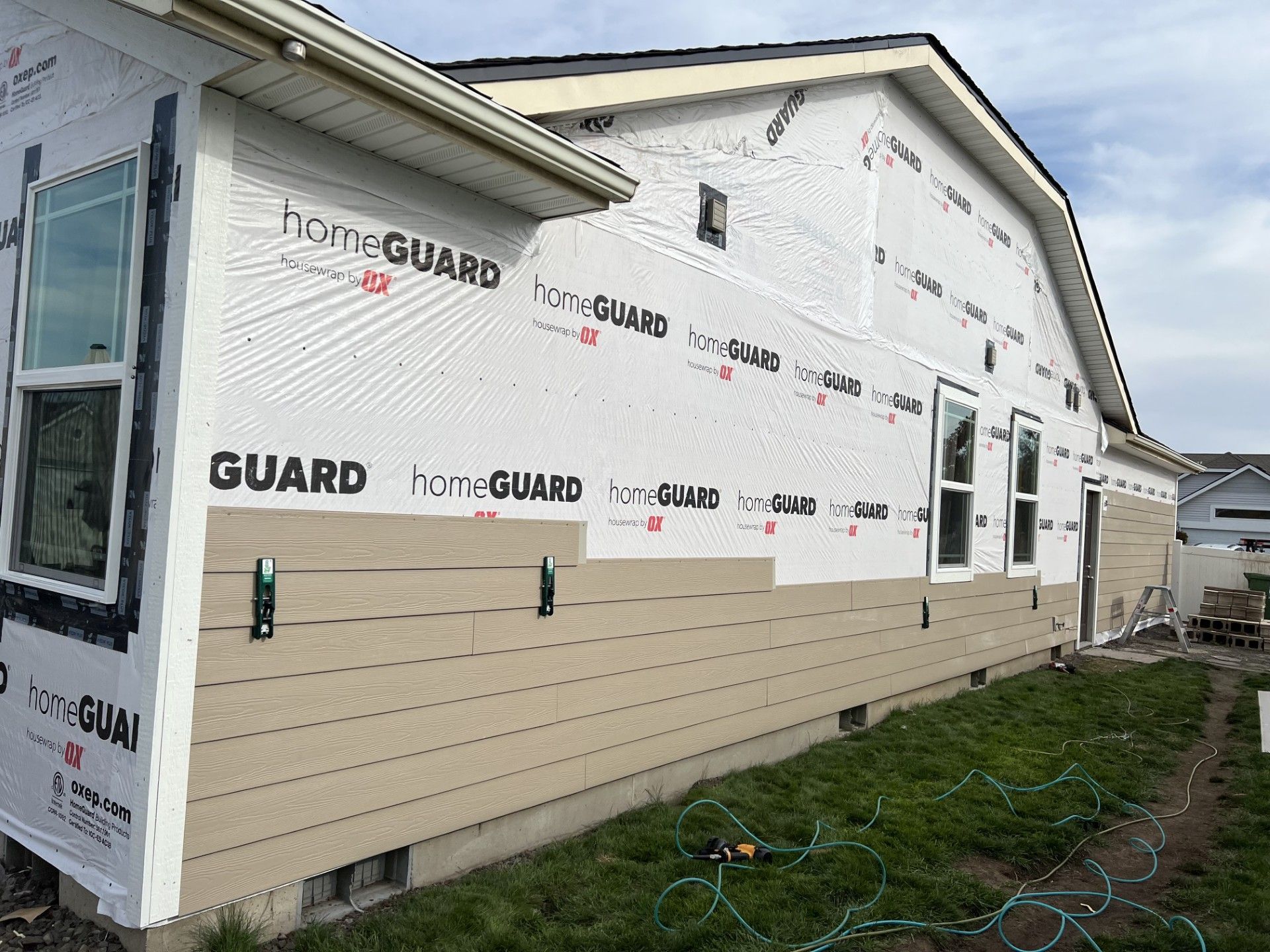 A house under construction with tan horizontal siding partially installed over a white protective house wrap.