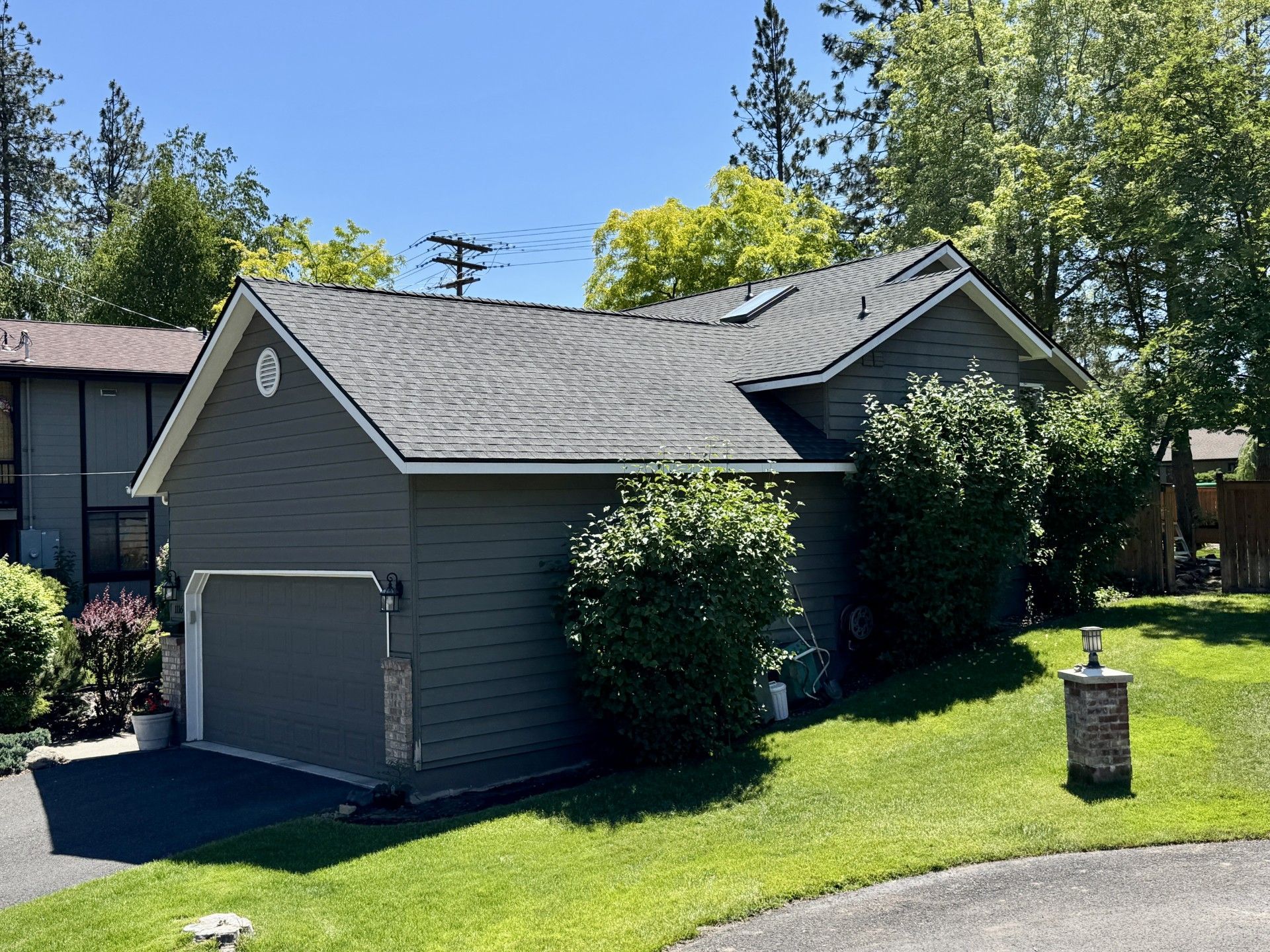 A gray, one-story house with a two-car garage, gray shingled roof, and green lawn on a sunny day.