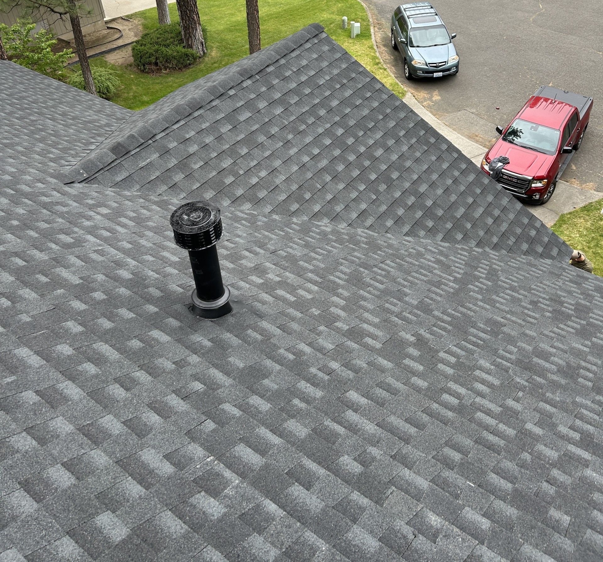 A high-angle view of a gray asphalt shingle roof with a black plumbing vent pipe, overlooking a driveway with two cars.