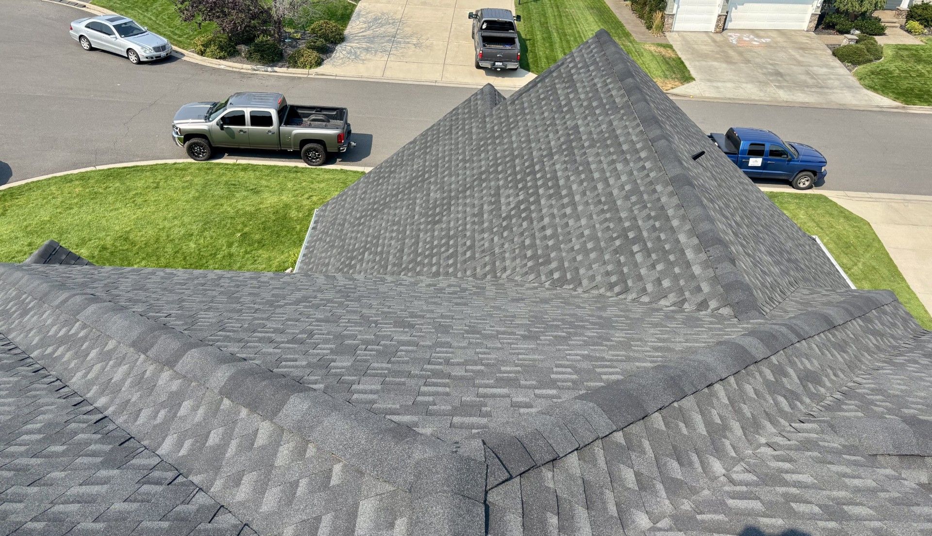 A high-angle view of a grey shingled roof with multiple peaks, overlooking a suburban street with parked vehicles.