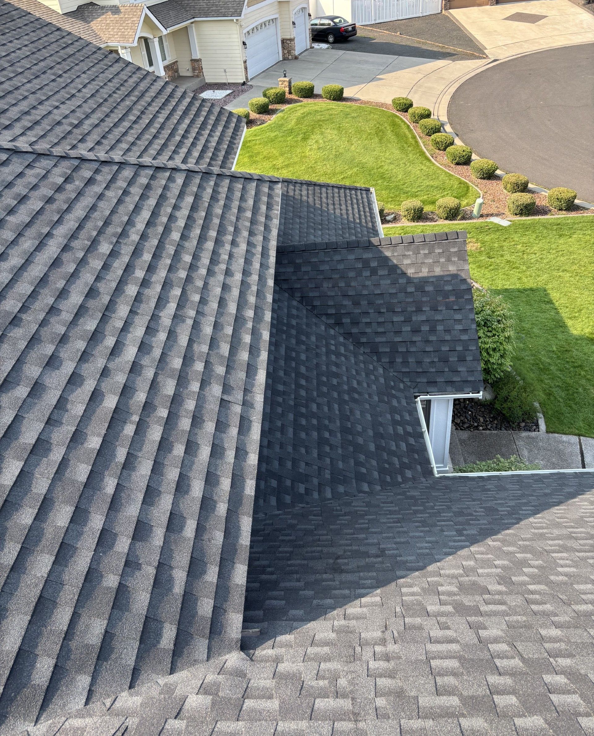 A high-angle view of a gray shingled residential roof with a valley and a glimpse of a lawn and driveway below.