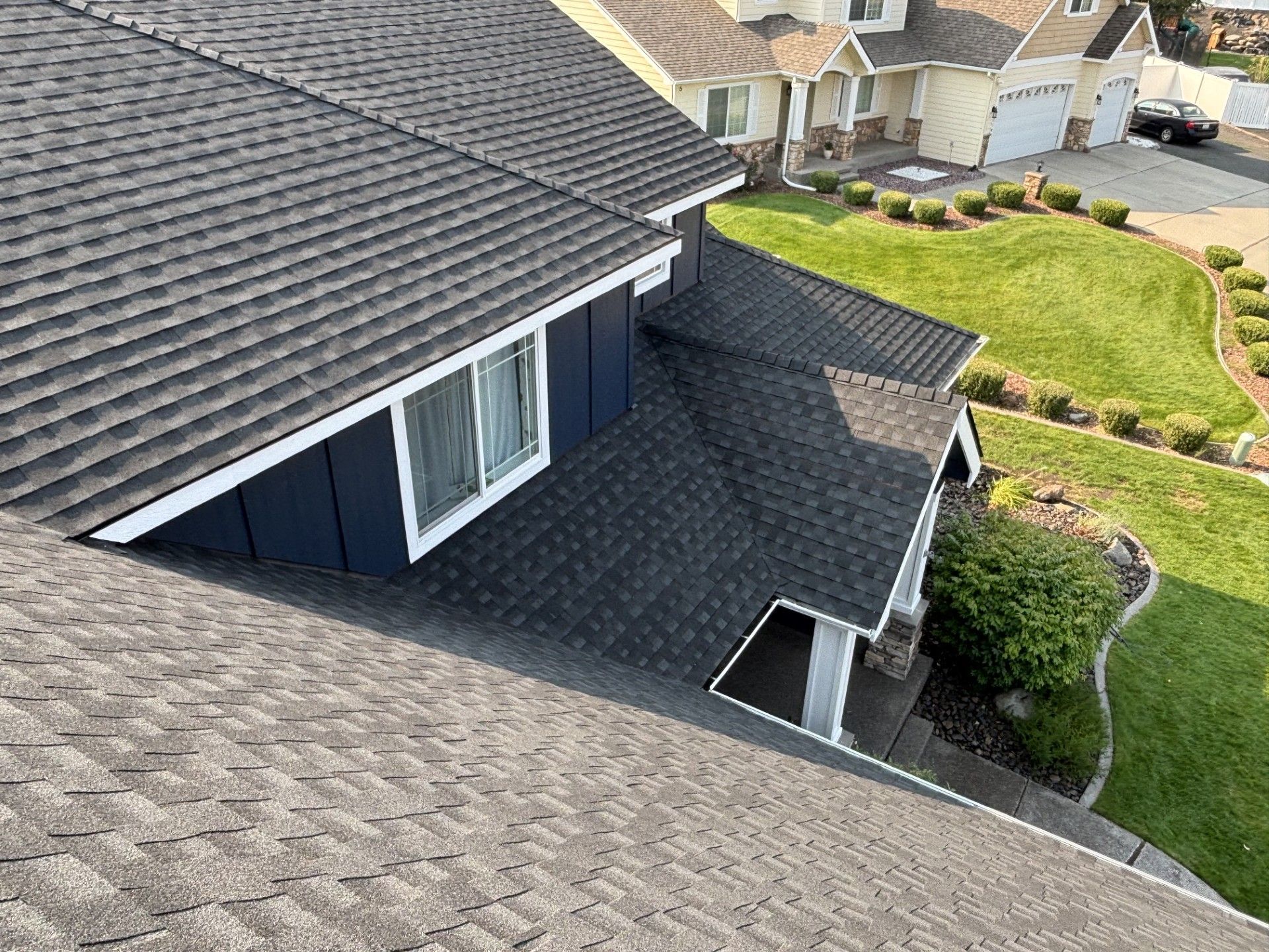 High-angle view of residential roofs with dark gray shingles, white trim, and a blue-sided dormer near a grassy lawn.