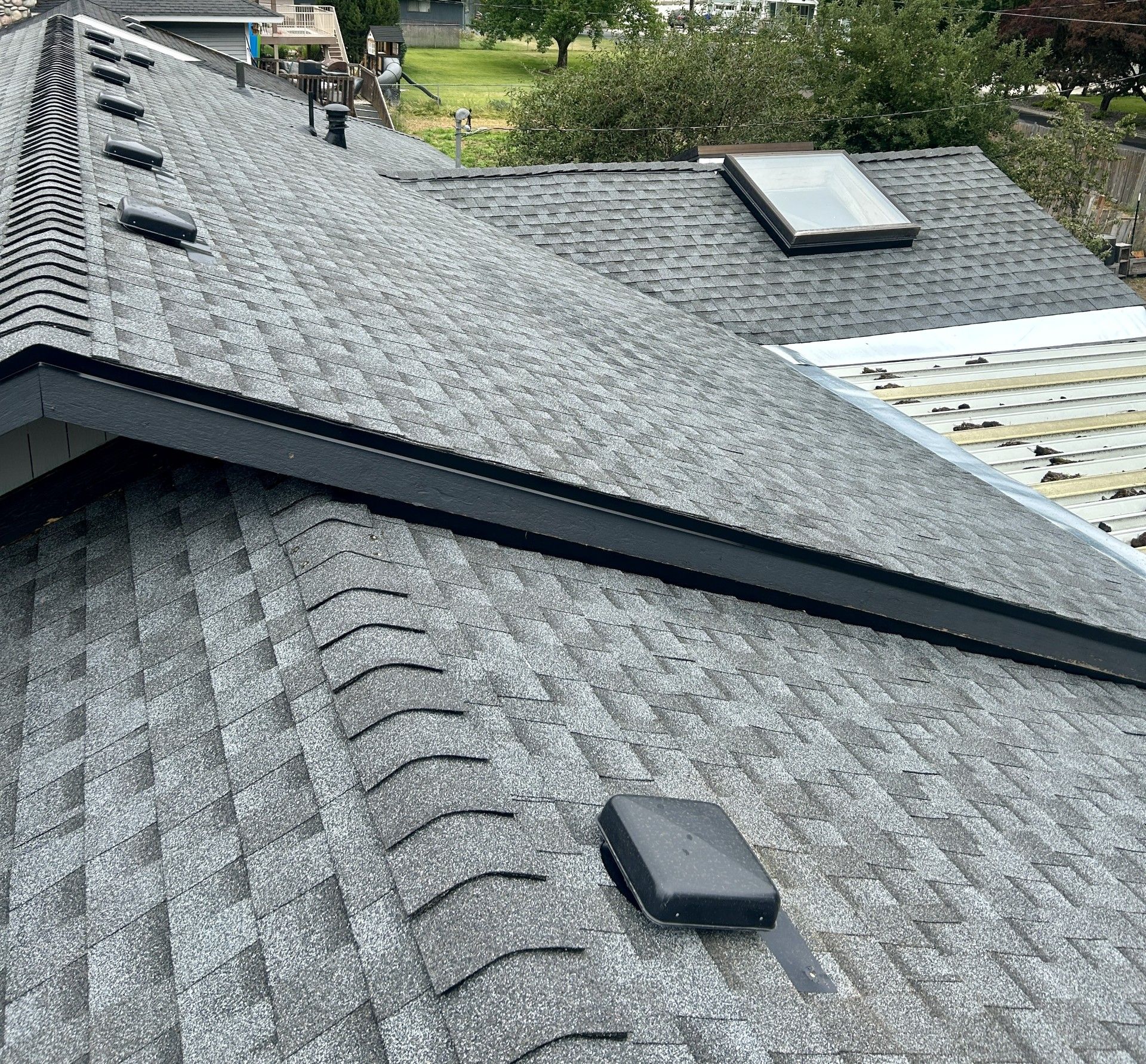 A high-angle view of a gray shingled roof with a skylight, vent caps, and a section of roof under construction.