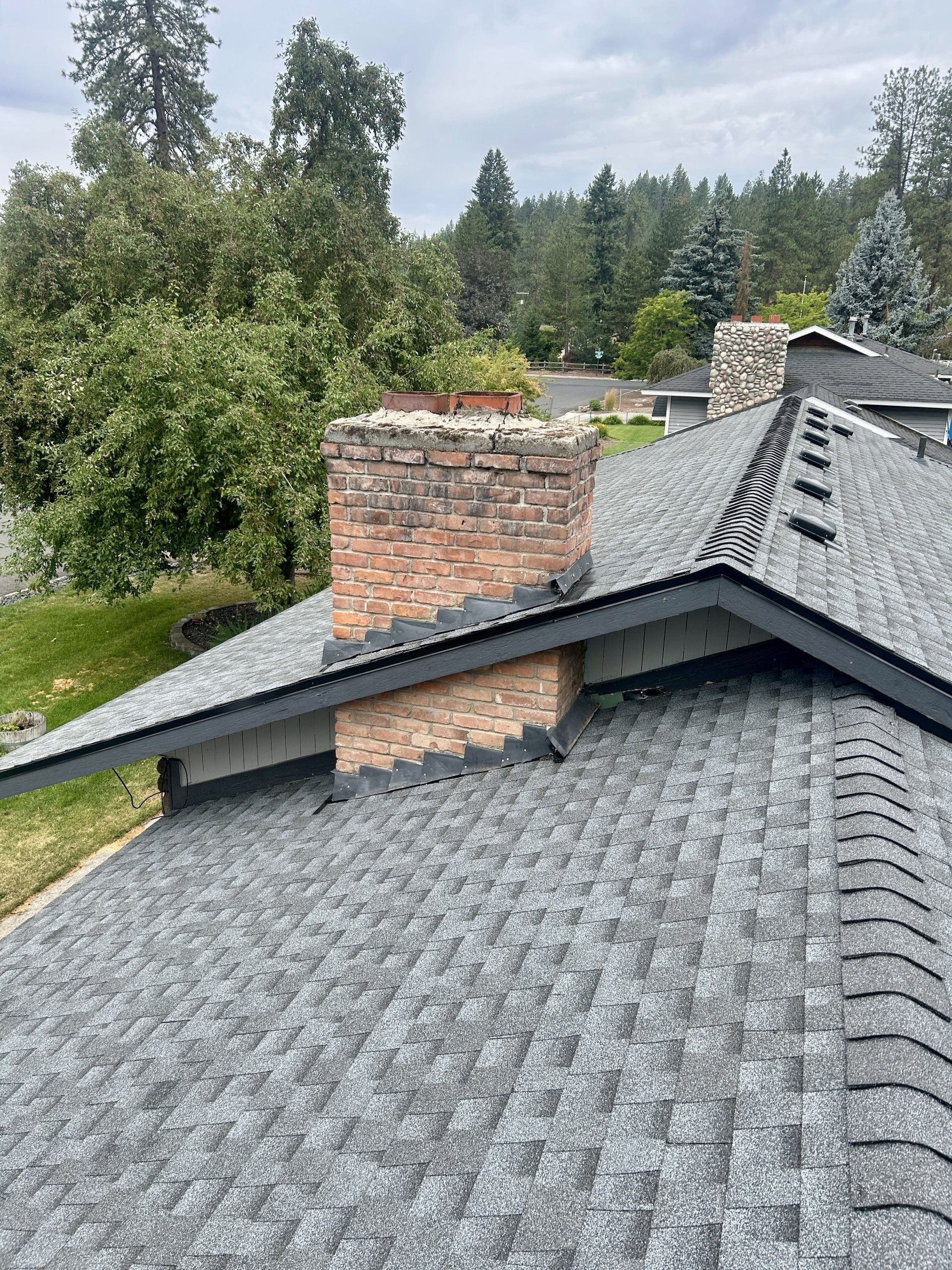 A high-angle view of a grey shingled roof with a brick chimney and dark trim, surrounded by trees under a cloudy sky.