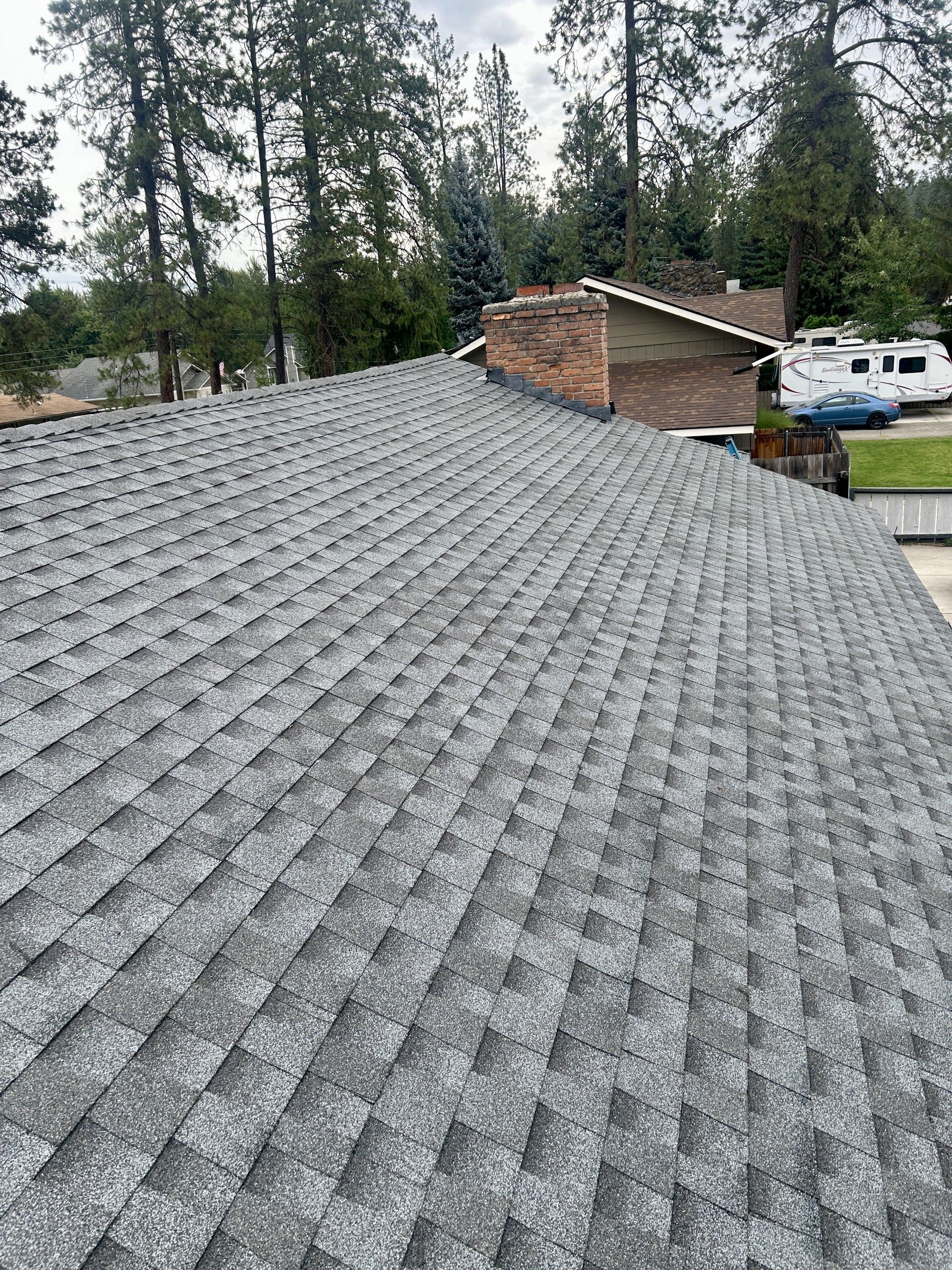A view looking down a residential gray asphalt shingle roof toward a brick chimney and treed background.